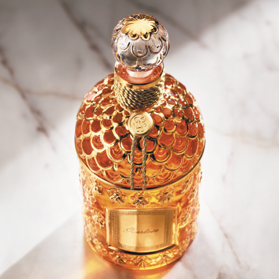 Um luxuoso e ornamentado frascos perfume com um design dourado e texturado e uma tampa elaborada, em contraste com uma superfície de mármore.