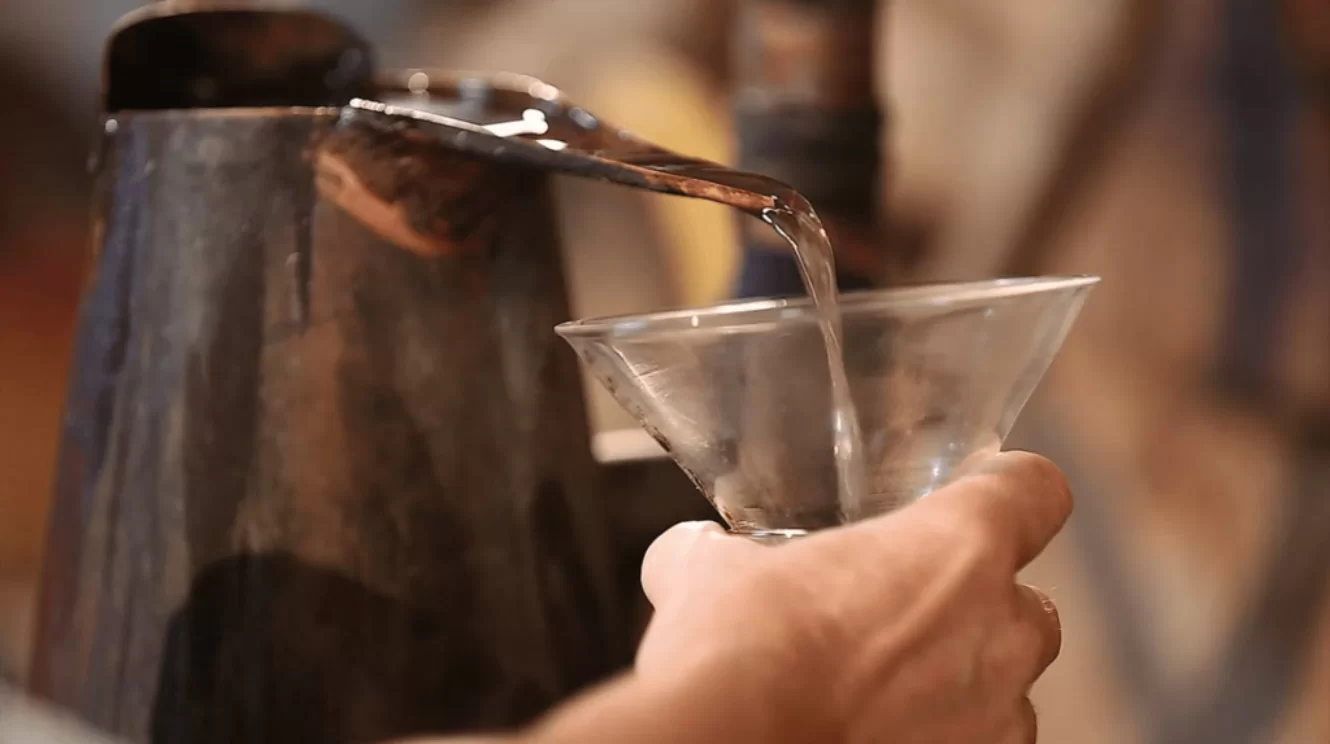Eine Person gießt eine klare Flüssigkeit aus einem Metallkessel in ein Glas. Der Hintergrund ist unscharf, sodass der Fokus auf dem Gießen liegt.