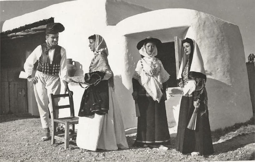 Una fotografia in bianco e nero che ritrae quattro persone in costume tradizionale in piedi davanti a un piccolo edificio in stile adobe. Due donne indossano gonne lunghe e scialli, mentre un uomo con gilet e cappello interagisce con loro.