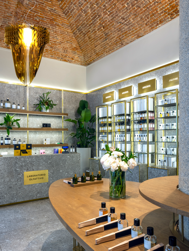 Un moderno negozio di profumi caratterizzato da un elegante bancone in legno con campioni di profumo e una vetrina con bottiglie disposte con eleganza sugli scaffali, impreziosito da piante verdi e un'elegante lampada a sospensione.