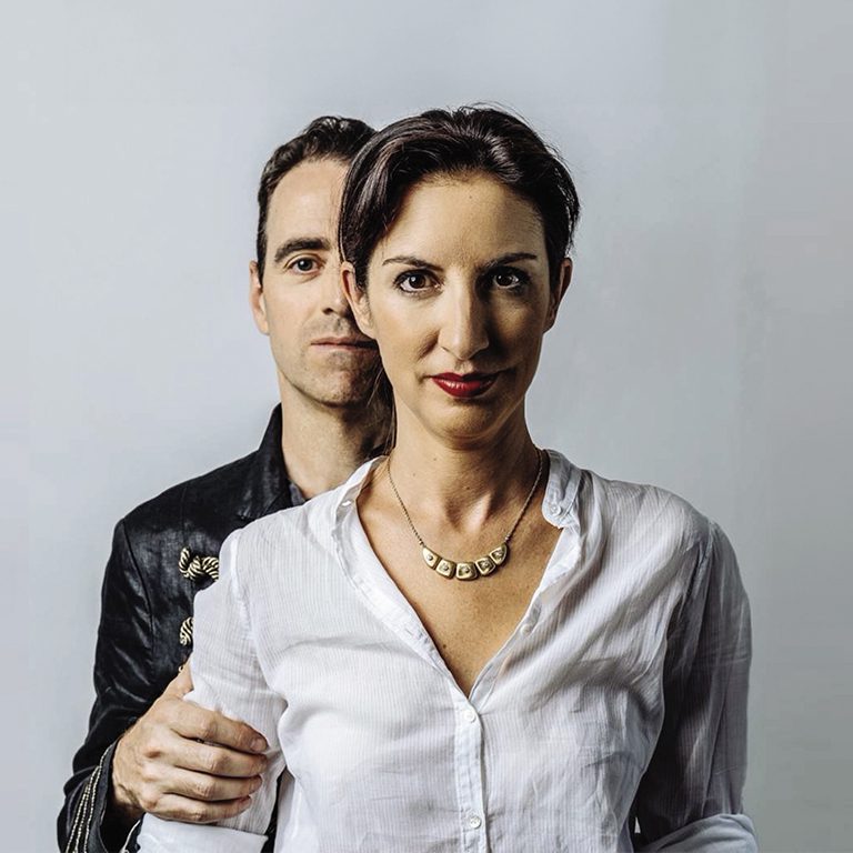 Un uomo è in piedi, parzialmente alle spalle di una donna, il cui sguardo è rivolto verso la fotocamera. La donna indossa una camicia bianca e una collana, mentre l'uomo ha indosso una giacca nera.