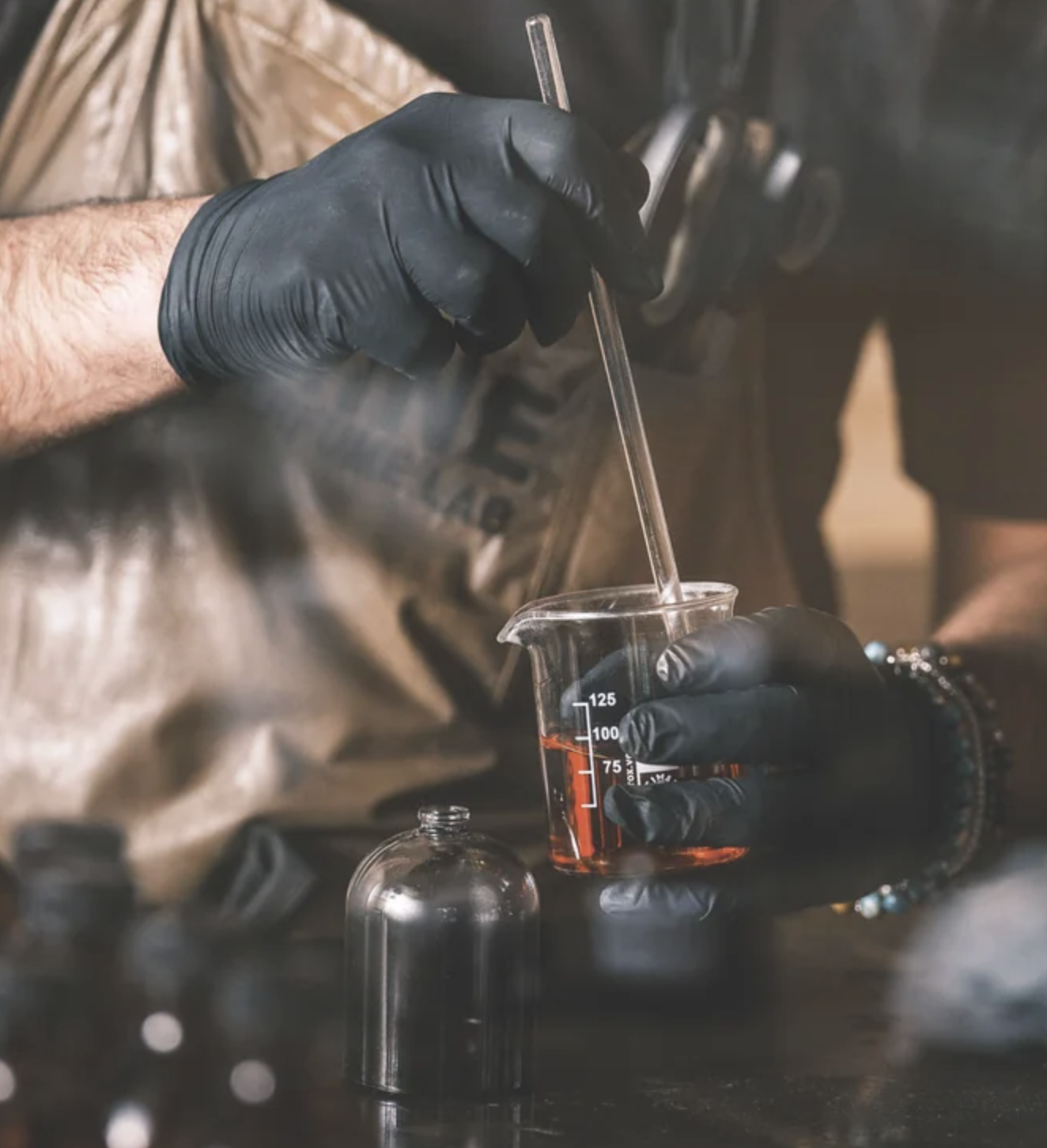 Uma pessoa usando luvas pretas está a mexer um líquido num copo com um agitador de vidro, tendo como pano de fundo um cenário desfocado de equipamentos de laboratório.