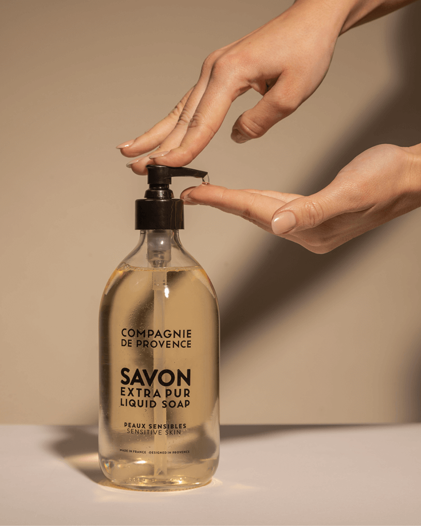 Una mano preme il dosatore di una bottiglia di sapone liquido "Savon Extra Pur", trasparente e posizionata su uno sfondo neutro. La bottiglia presenta un'etichetta nera con il marchio "Compagnie de Provence".