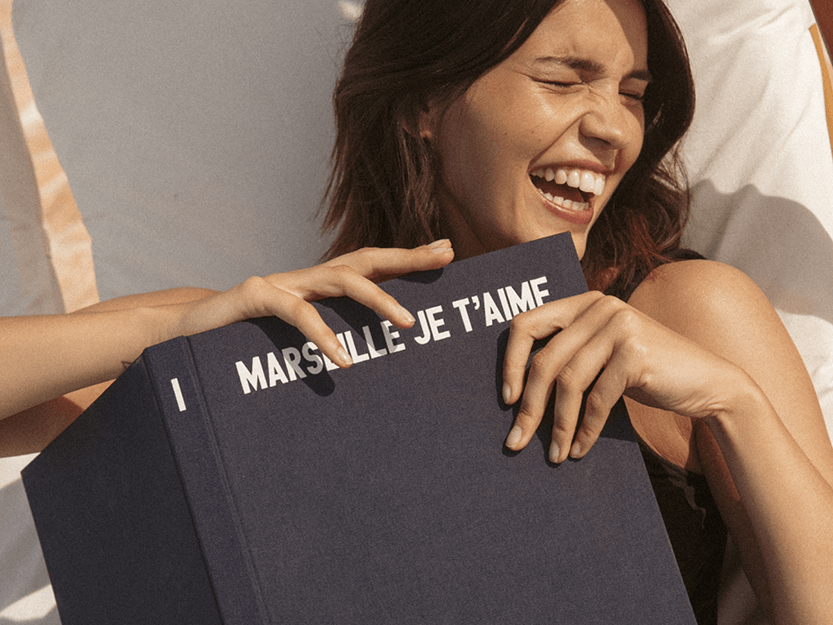 Una donna sorride gioiosamente mentre tiene in mano un grande libro intitolato "Marsaille Je T'aime", sdraiata su un lettino.
