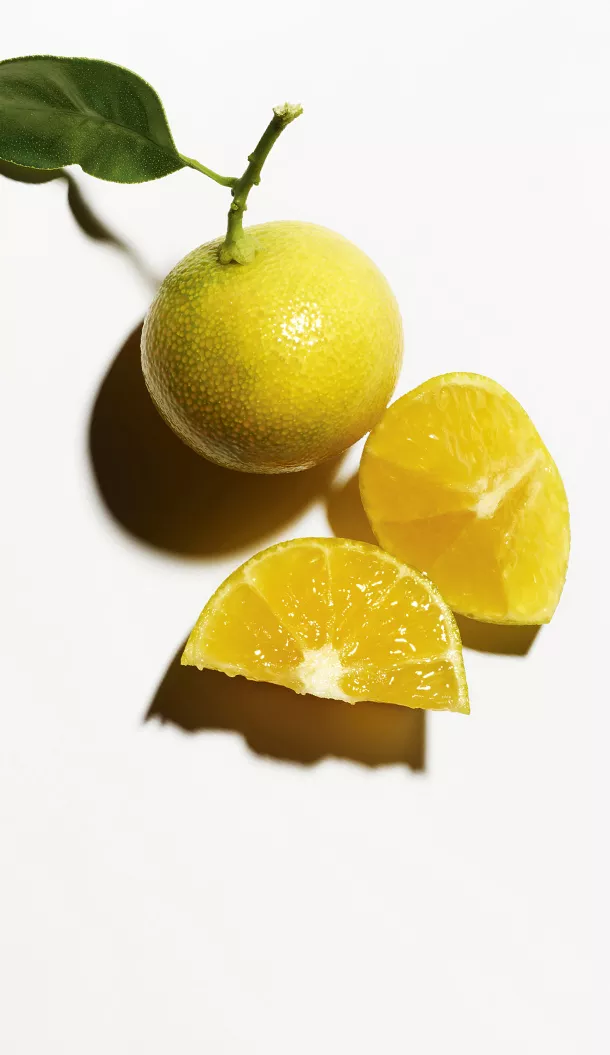 A whole yellow citrus fruit with a green leaf and two freshly cut slices, all resting on a white surface.