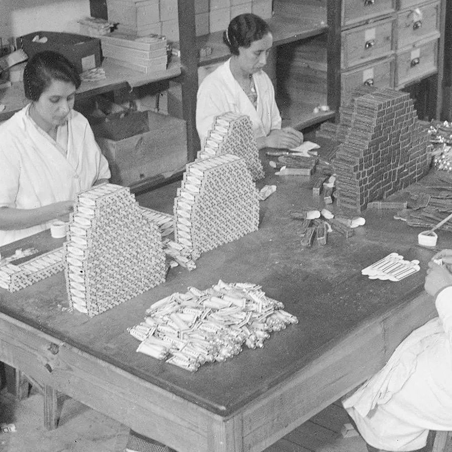 Duas mulheres estão sentadas à mesa de madeira a empacotar produtos empilhados, com vários artigos e materiais cuidadosamente dispostos à sua volta, num ambiente de oficina.