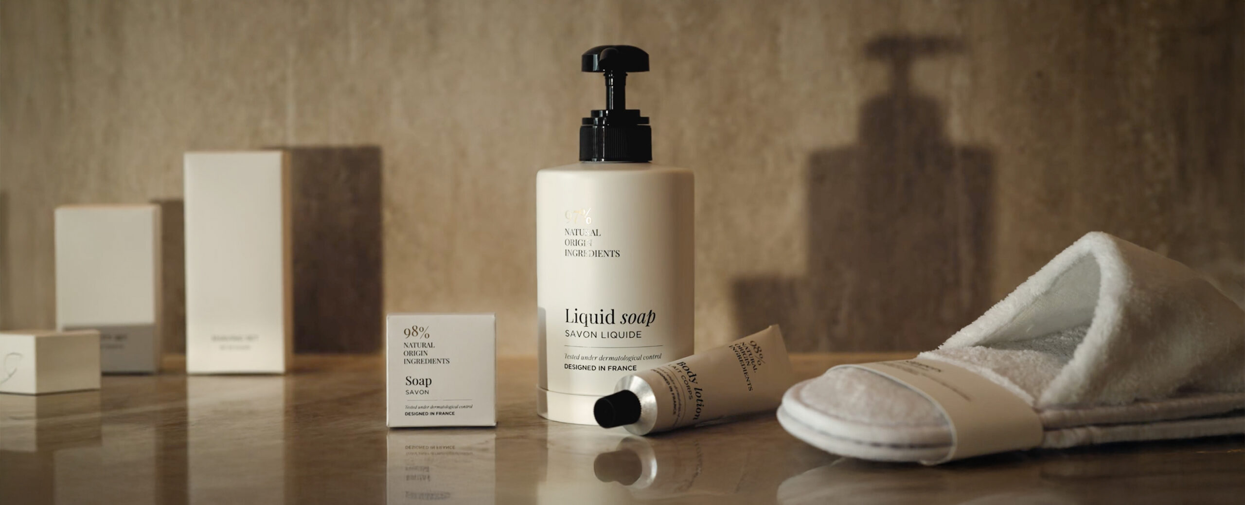 Um frasco de sabonete líquido é exibido sobre uma superfície de madeira ao lado de uma pequena caixa de sabonete, um tubo de loção e um chinelo branco felpudo, tudo contra um fundo neutro.