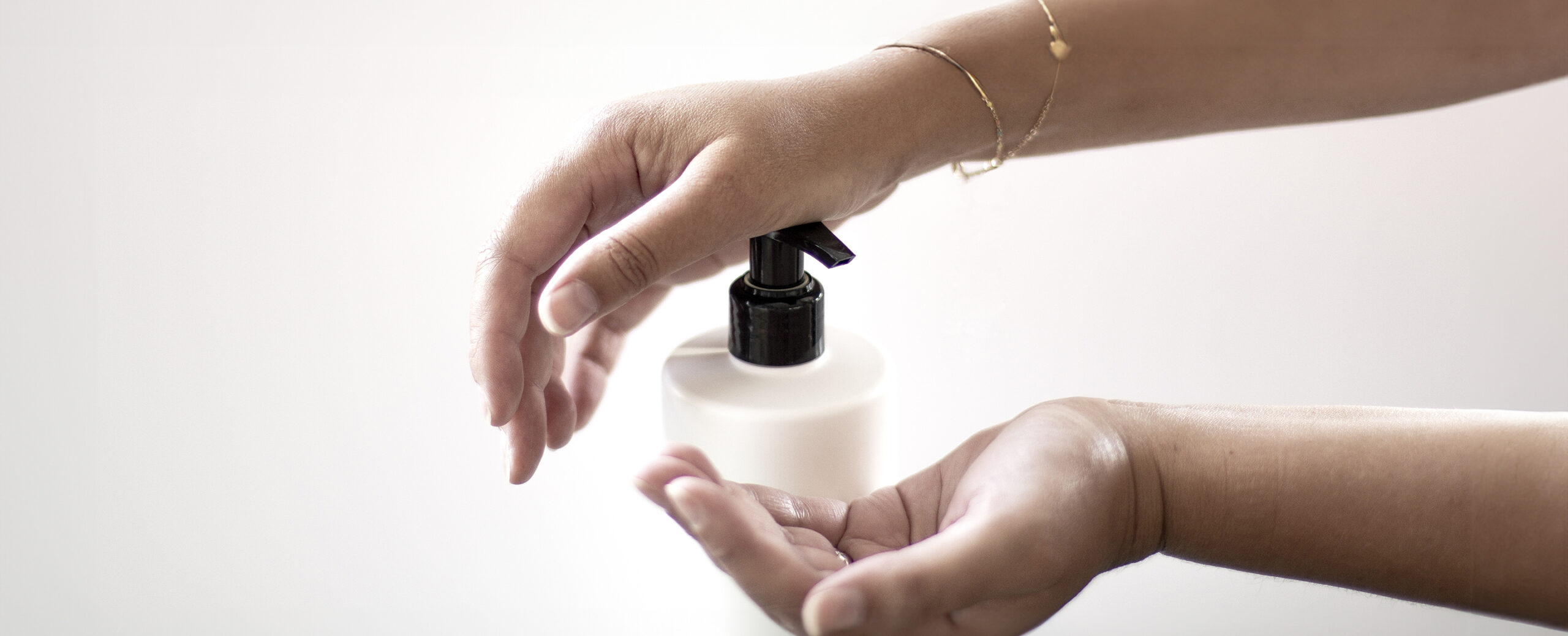 As mãos de uma pessoa são mostradas no processo de dispensar líquido de um frasco com bomba branca para a palma da mão. O fundo é suave e neutro, com foco nas mãos e no frasco.