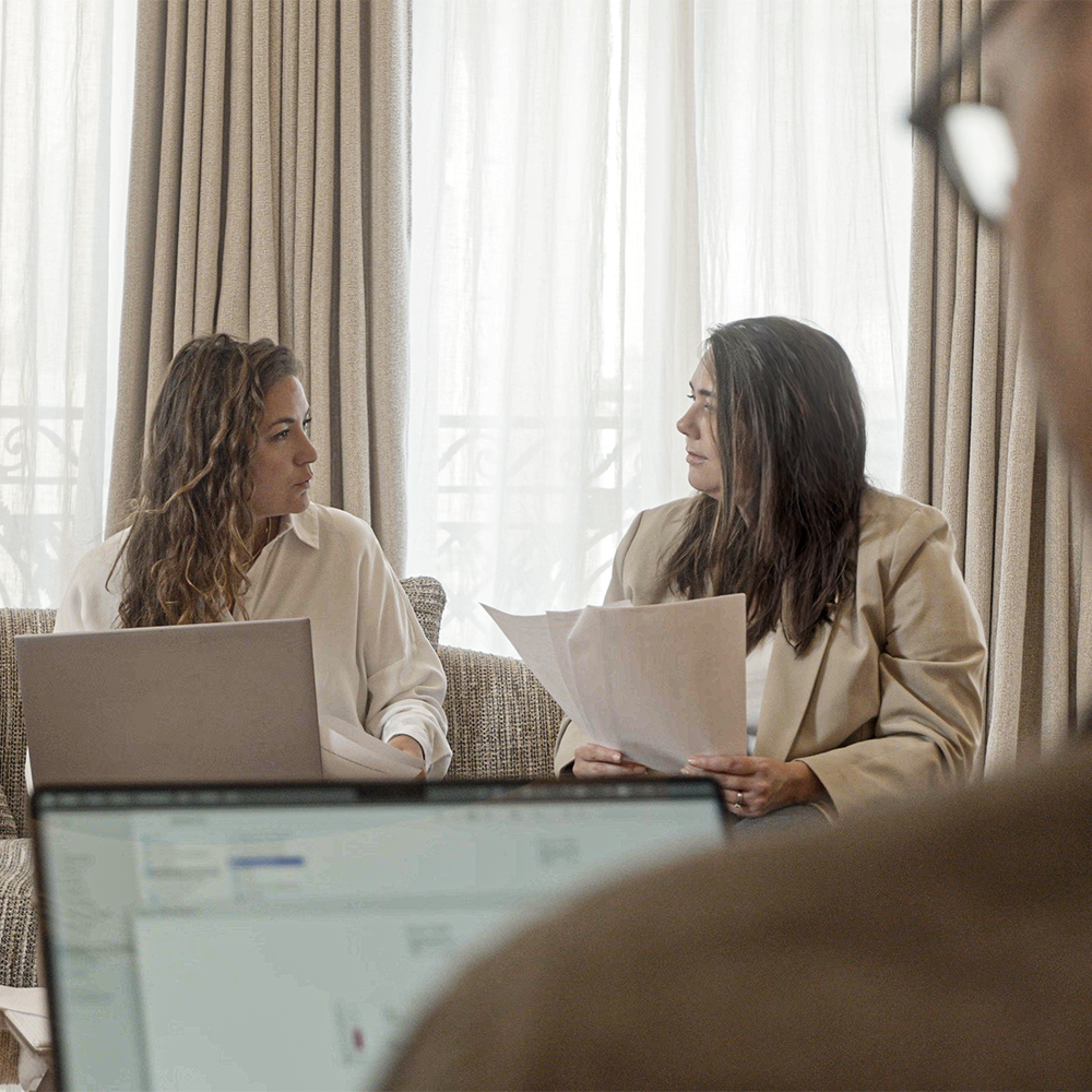 Duas mulheres estão sentadas numa sala bem iluminada, envolvidas numa discussão enquanto seguram papéis. Um computador portátil é visível em primeiro plano, sugerindo um ambiente de trabalho colaborativo.