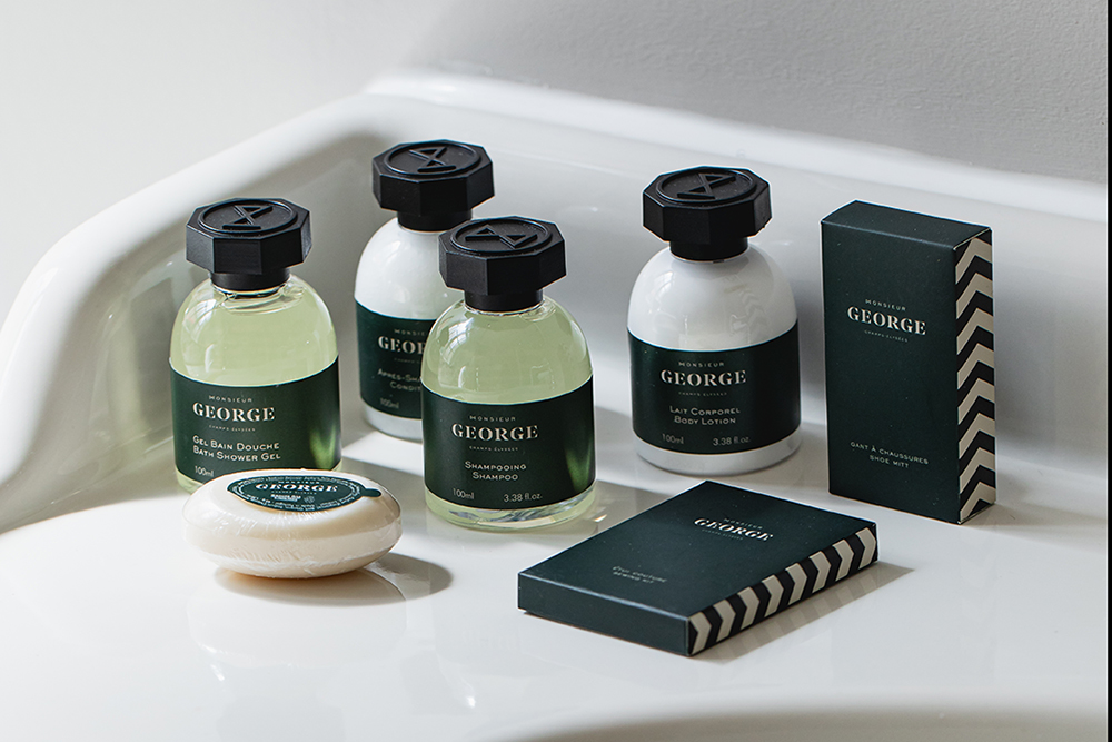 Uma coleção de produtos de higiene pessoal, incluindo champô, condicionador e sabonete, todos com a marca «George» e embalagens verdes e pretas, expostos numa bancada branca.