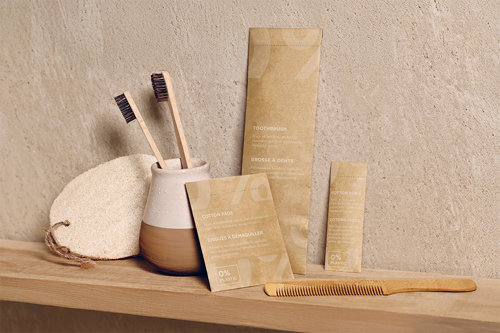Uma prateleira de madeira exibe artigos de higiene pessoal ecológicos: duas escovas de dentes de bambu, almofadas de algodão biodegradáveis, um pente de madeira suave e uma bucha natural, todos em embalagens minimalistas contra uma parede texturizada.