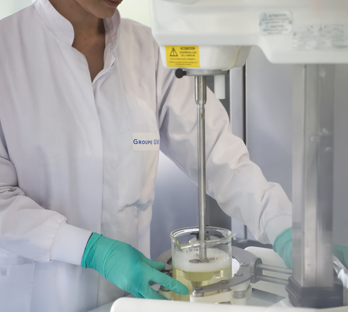 Um técnico de laboratório com bata branca e luvas verdes está a usar um misturador mecânico para mexer um líquido transparente num copo de vidro. A máquina faz parte de um equipamento científico.