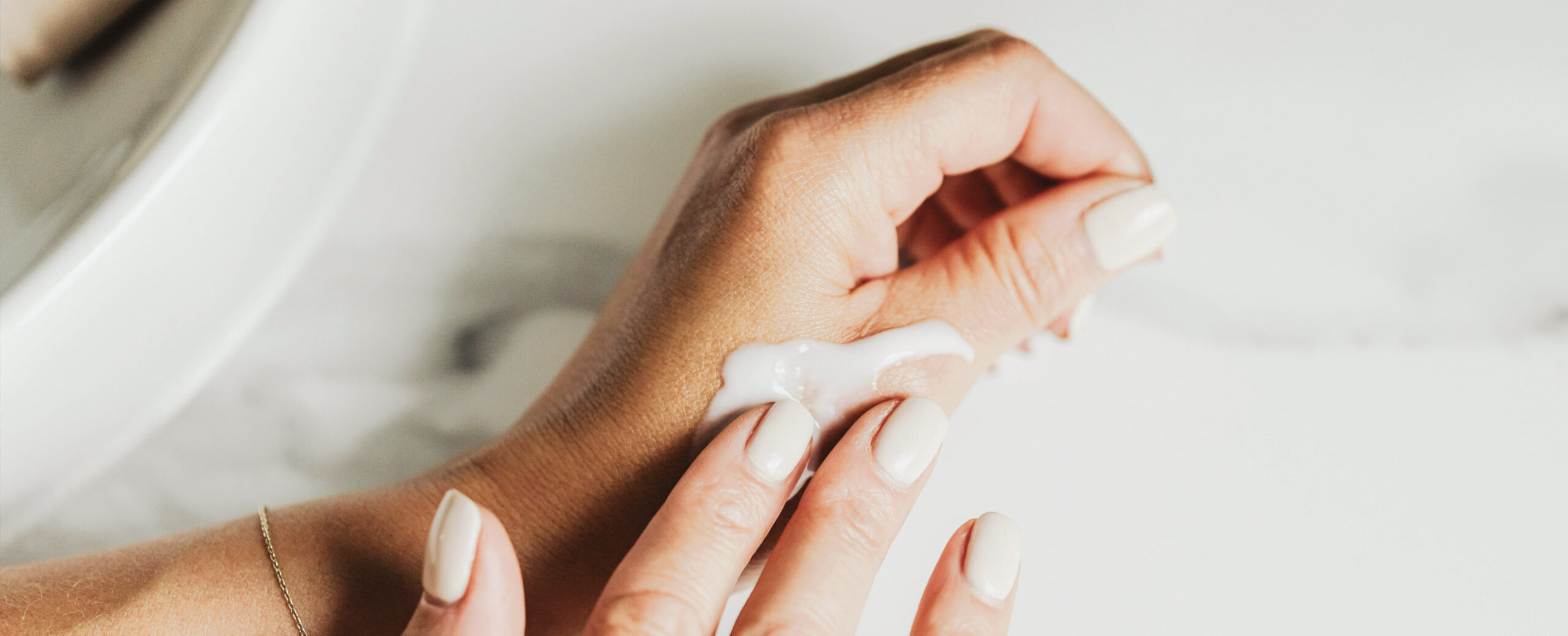 Close-up de uma mão a aplicar um creme branco no pulso, sobre uma superfície de mármore claro. As unhas estão bem cuidadas, com uma cor bege suave.