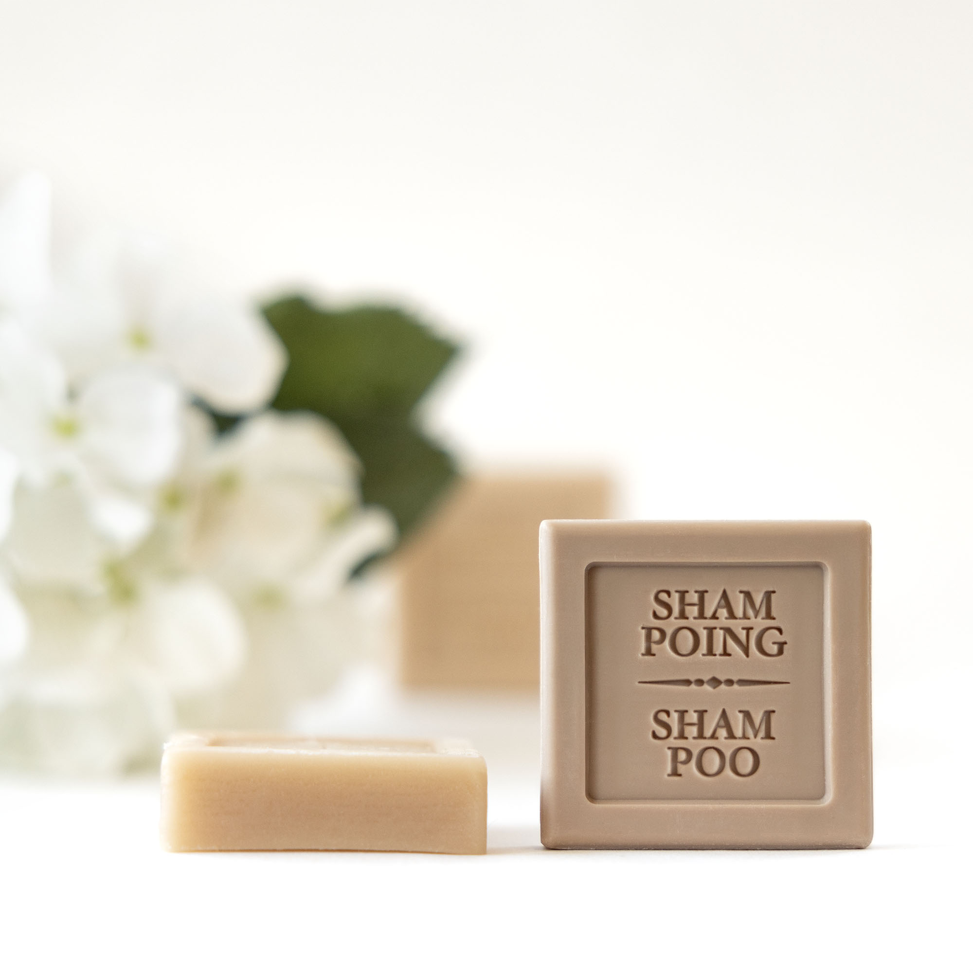 Ein Stück Shampoo-Seife mit der Aufschrift „SHAMPOOING“ neben einem kleineren rechteckigen Stück, mit zarten weißen Blüten und grünen Blättern im Hintergrund.