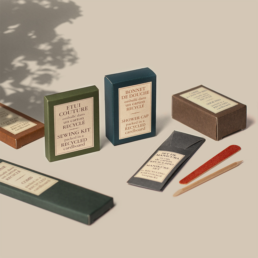 Una collezione di scatole di imballaggio ecologiche per vari prodotti, tra cui un kit da cucito e una cuffia da doccia, disposte artisticamente con bastoncini di legno e un'ombra sullo sfondo. Le scatole sono realizzate in cartone riciclato e presentano etichette minimaliste.