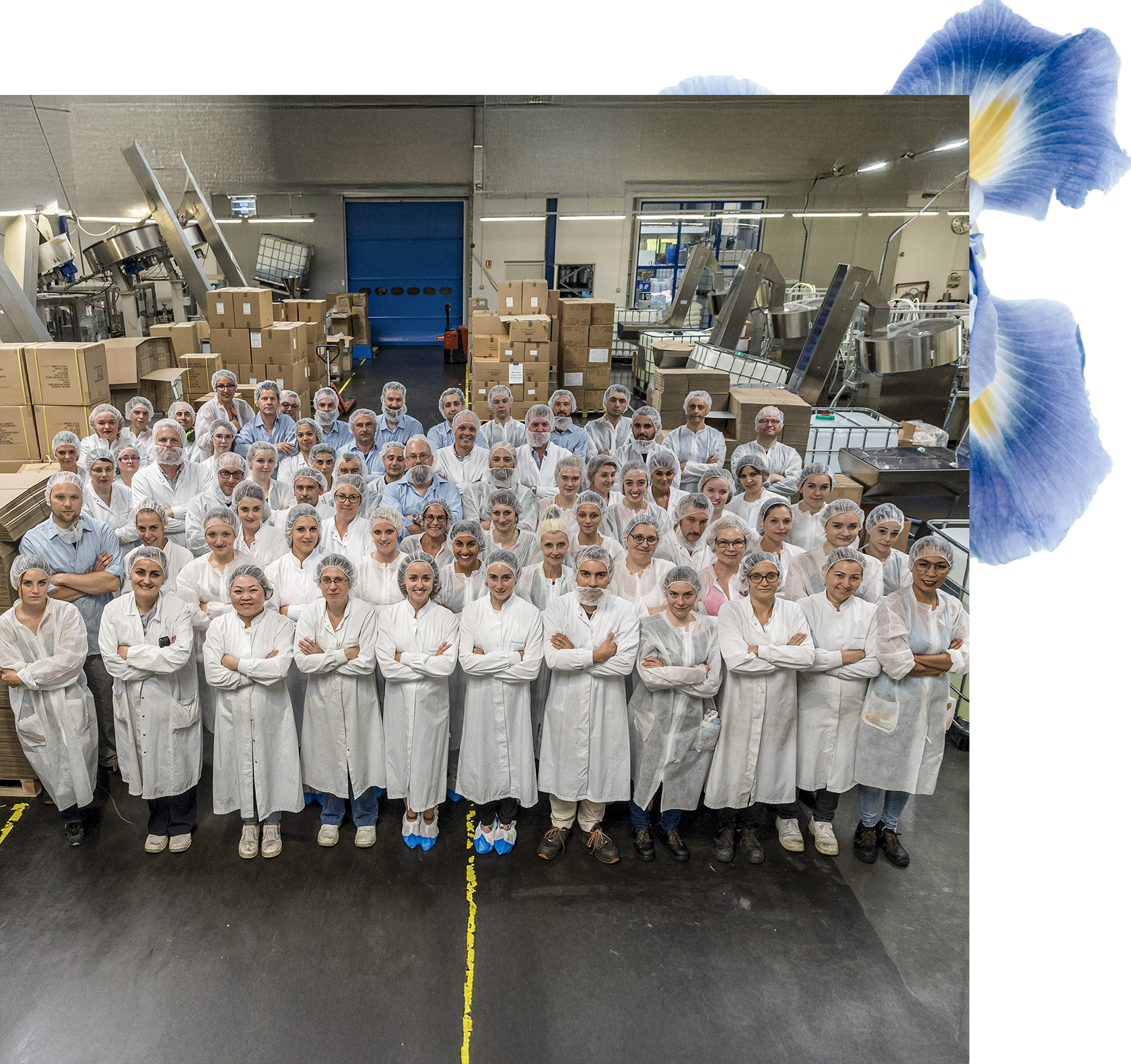 Un folto gruppo di oltre 50 persone in camice bianco e cuffia sta in piedi in una fabbrica, sorridendo alla telecamera con le braccia incrociate. Sullo sfondo si vedono scatole e macchinari.