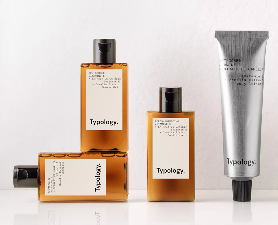 Drei Typology werden präsentiert: ein Duschgel und eine Shampooflasche, beide in minimalistischem Design, sowie eine Tube Körperlotion, alle mit eleganten Etiketten, auf denen Kamelienextrakt im Mittelpunkt steht.