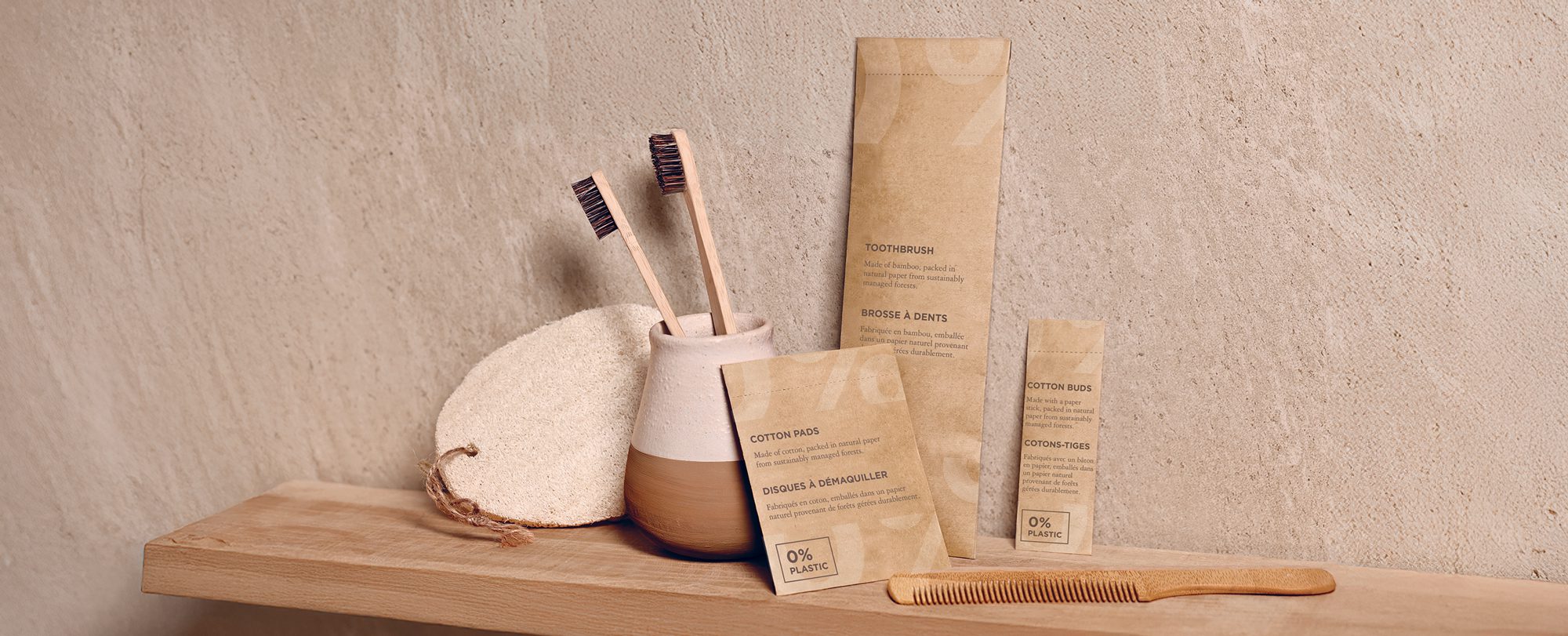 Produtos ecológicos para casa de banho dispostos numa prateleira de madeira, incluindo escovas de dentes de bambu, um pente de madeira e embalagens biodegradáveis para cotonetes e almofadas de algodão. O fundo apresenta uma parede texturizada para uma estética natural.