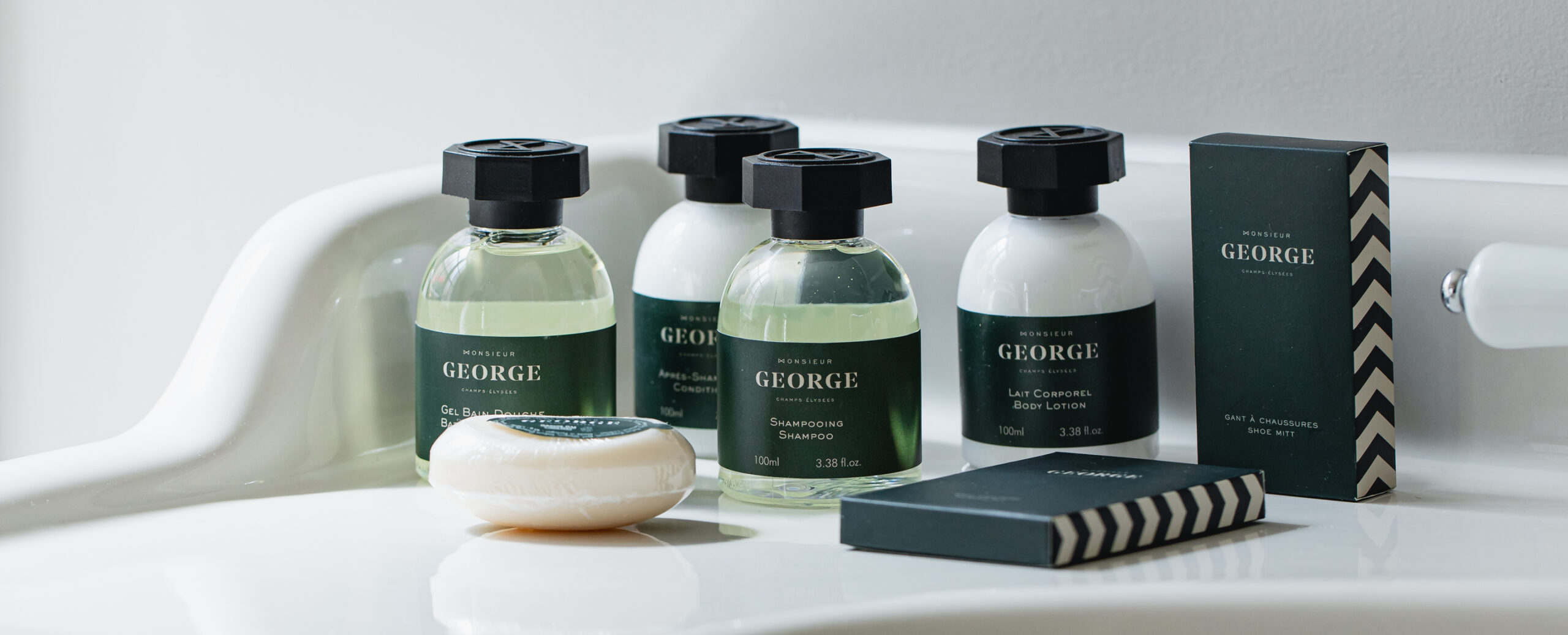 Uma coleção de produtos de higiene pessoal sobre uma pia branca, incluindo champô, loção corporal, um sabonete e uma caixa, todos com a etiqueta «George». Os frascos têm um design elegante com tampas pretas.