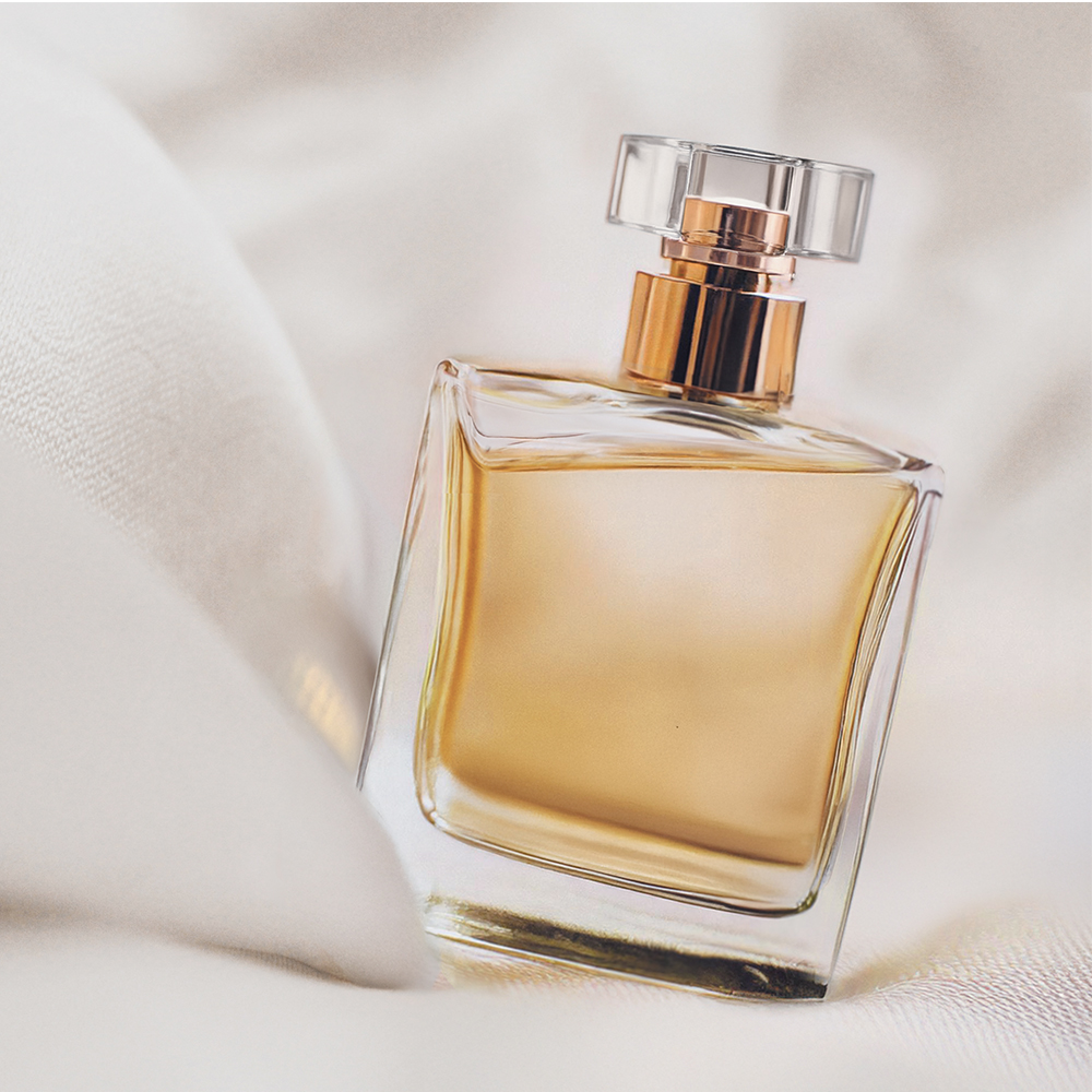 Um frasco de perfume de vidro transparente com um líquido dourado no interior, colocado sobre um fundo de tecido branco suave. O frasco apresenta um design simples e elegante com uma tampa redonda.
