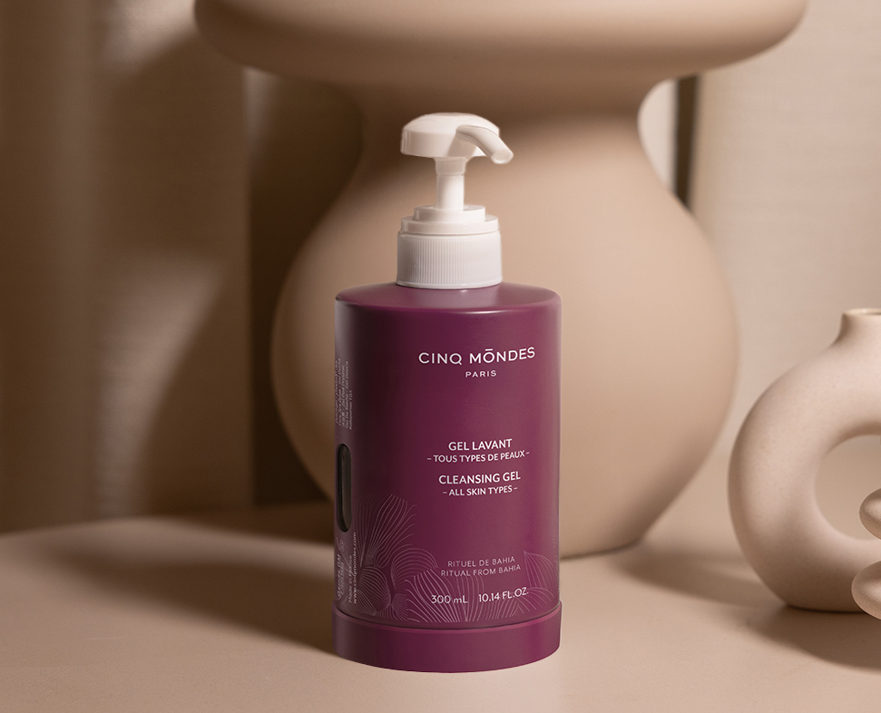 Frasco de gel de limpeza roxo com dispensador em bomba, com a etiquetaCinq Mondes » e indicado para todos os tipos de pele, apresentado contra um fundo neutro.