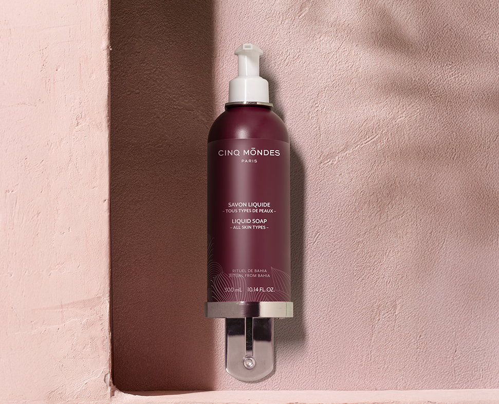 Um frasco de sabonete líquido da Cinq Mondes , com um dispensador de bomba, apoiado numa parede rosa texturada. O frasco tem um design elegante, com um rótulo roxo escuro e fundo transparente.