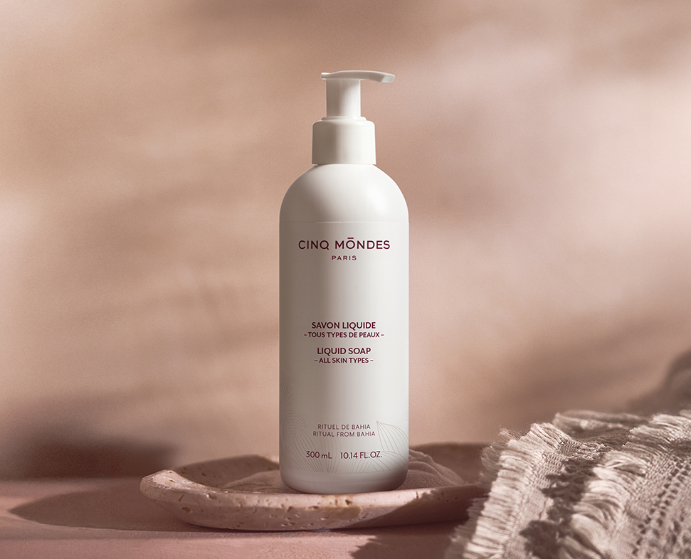 Um frasco de sabonete líquido da Cinq Mondes , com a etiqueta «Savon Liquide - Tous Types de Peaux» (Sabonete Líquido - Todos os Tipos de Pele), repousa sobre uma superfície texturada ao lado de um prato raso. O fundo apresenta cores suaves e discretas, realçando a apresentação elegante do produto.