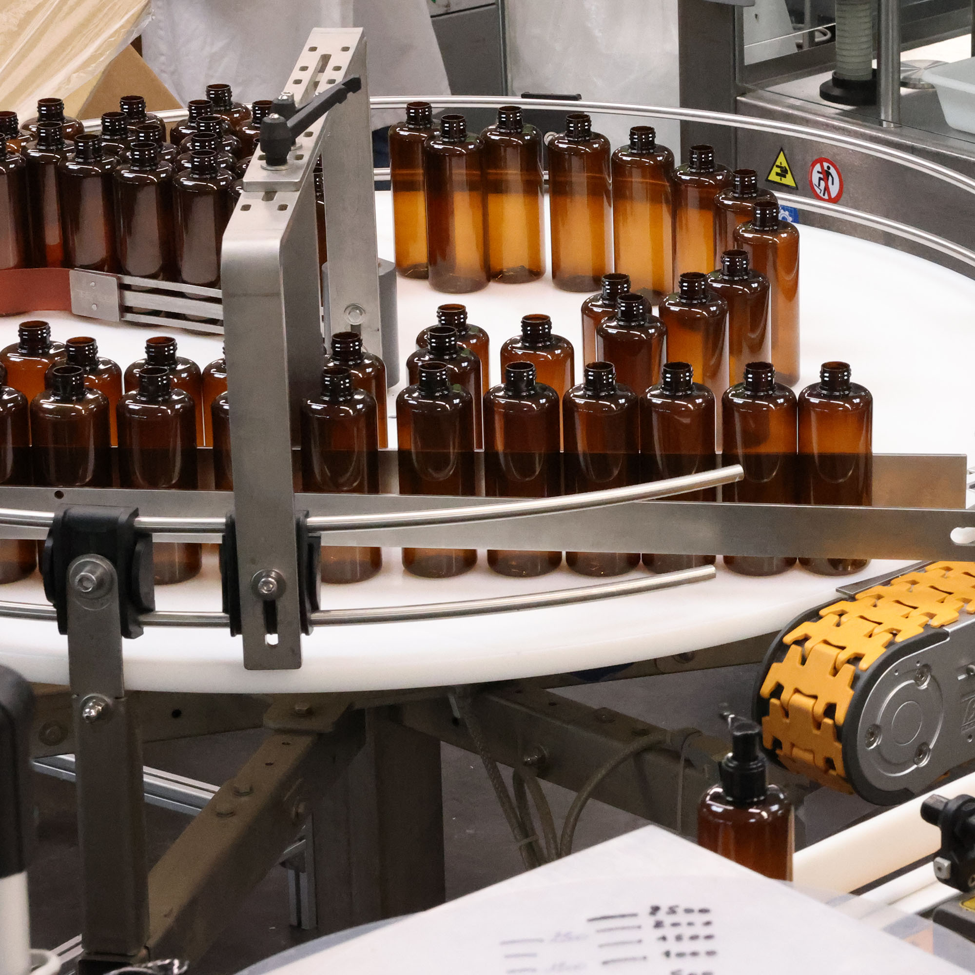 Uma correia transportadora circular numa fábrica exibe filas de garrafas de vidro castanho, refletindo um ambiente de produção movimentado. Ferramentas e equipamentos são visíveis ao fundo.