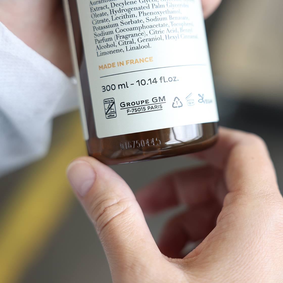 Uma pessoa segurando um frasco castanho com uma lista detalhada de ingredientes e o rótulo «Fabricado em França», com foco na parte de trás do frasco. O volume do frasco é de 300 ml.