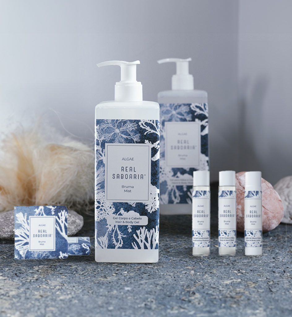 Una collezione di prodotti per la cura della pelle, tra cui un flacone grande di spray e tubetti più piccoli, caratterizzati da un disegno blu e bianco a forma di corallo su uno sfondo neutro. Sono visibili anche pietre decorative e texture morbide.