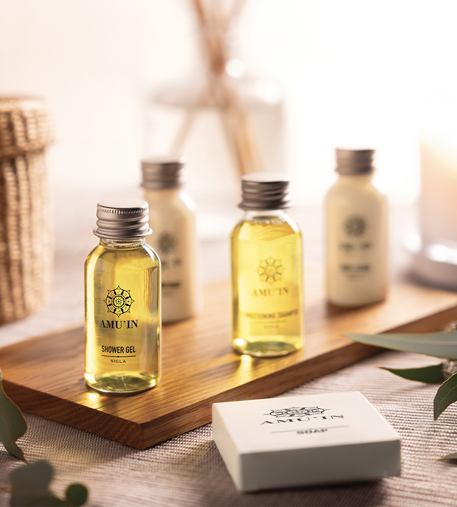 Una collezione di flaconcini contenenti gel doccia e shampoo, disposti su un vassoio di legno con uno sfondo in tessuto naturale ed elementi decorativi.