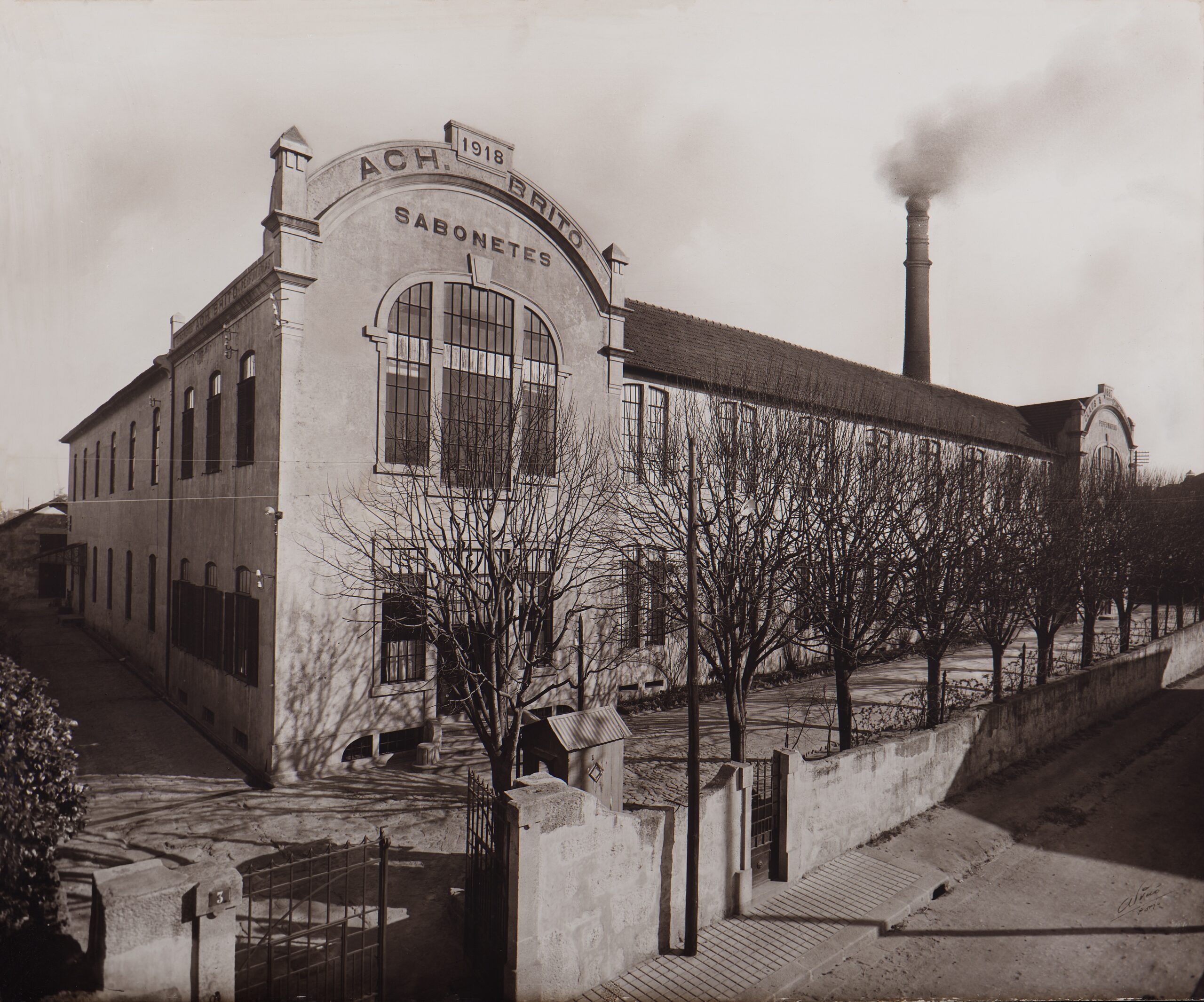 Storico edificio industriale con l'anno 1918 riportato sulla facciata, caratterizzato da un'alta ciminiera da cui fuoriesce del fumo e circondato da alberi spogli.