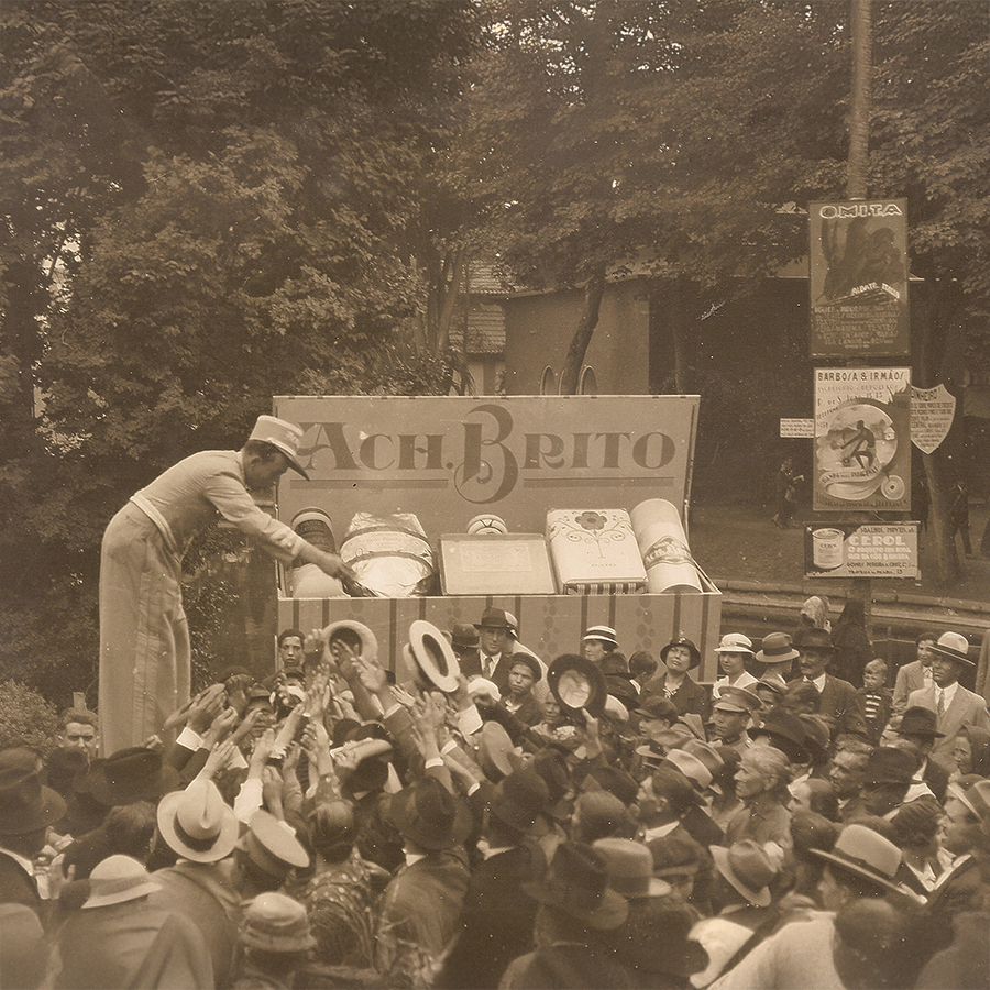 Una foto d'epoca che ritrae una folla di persone protese verso un venditore di alta statura che distribuisce prodotti da un grande cartellone su cui si legge "A. C. H. Brito". La scena è animata da cappelli e volti entusiasti, a testimonianza di un evento vivace.