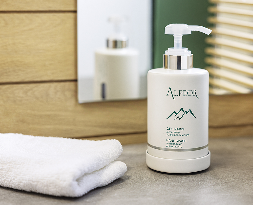 Um frasco de gel Alpeor está sobre uma bancada ao lado de uma toalha branca dobrada, com um espelho e painéis de madeira ao fundo.