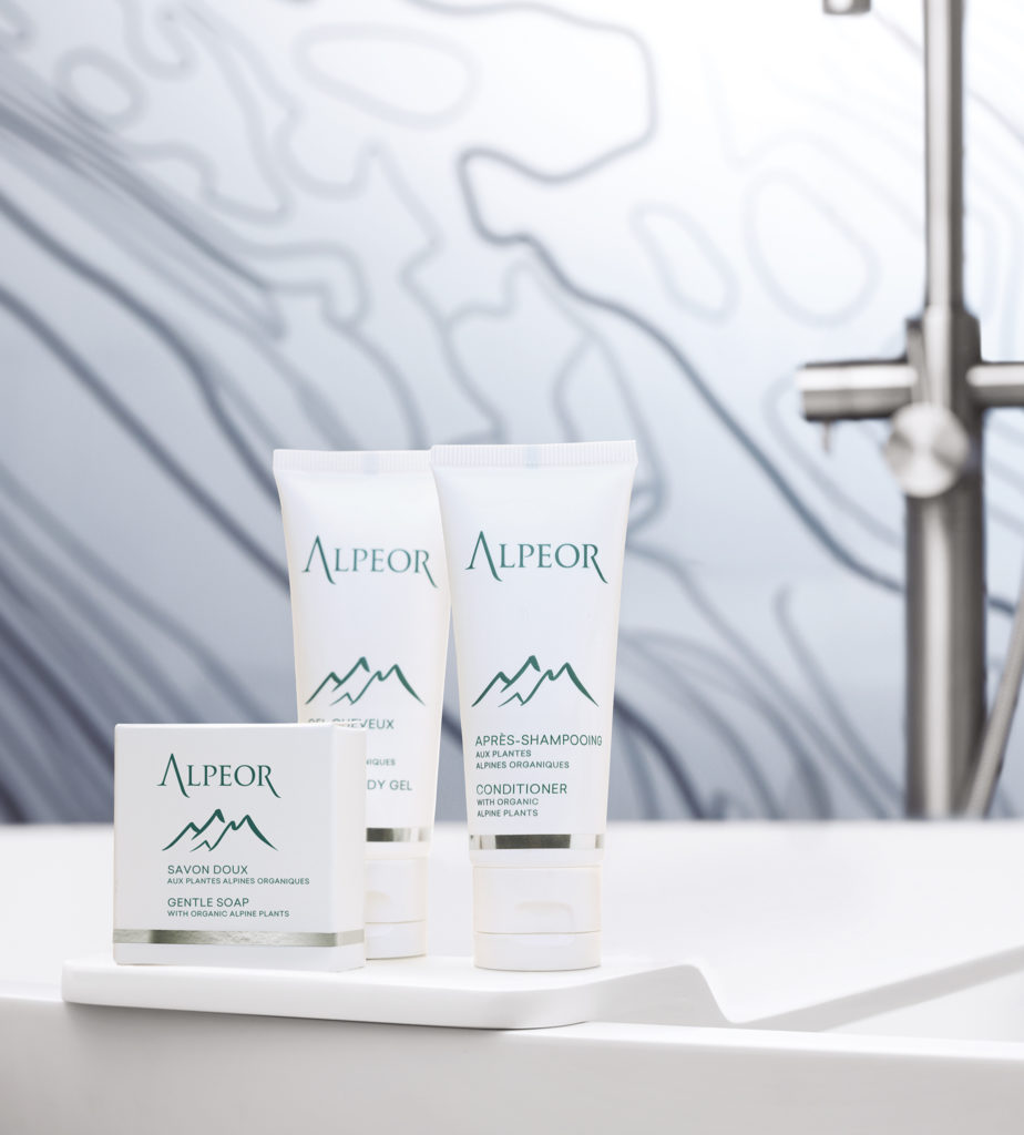 Três produtos Alpeor , incluindo um sabonete suave, um gel corporal e um condicionador, expostos sobre uma superfície branca contra um fundo com um padrão suave.