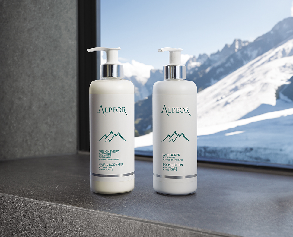 Duas garrafas de gel Alpeor e corpo e loção corporal Alpeor estão expostas numa bancada de pedra, com uma vista panorâmica das montanhas cobertas de neve visíveis através de uma grande janela.