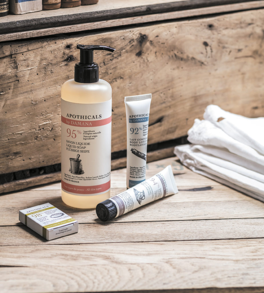 Uma seleção de produtos de farmácia dispostos sobre uma superfície de madeira, incluindo um frasco grande de sabonete líquido, uma loção hidratante, um creme para as mãos e um sabonete em barra. Toalhas de tecido macio são parcialmente visíveis ao fundo.