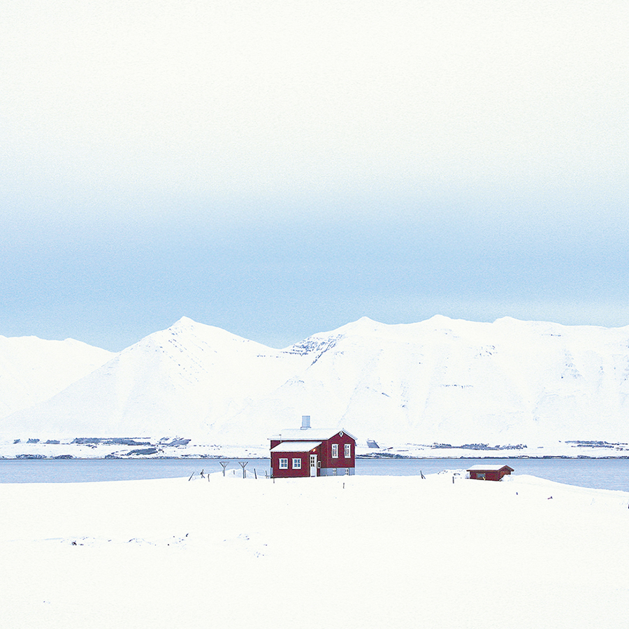 Ein kleines rotes Haus steht einsam in einer verschneiten Landschaft, umgeben von weißen Bergen und einem ruhigen Gewässer unter einem blassblauen Himmel.