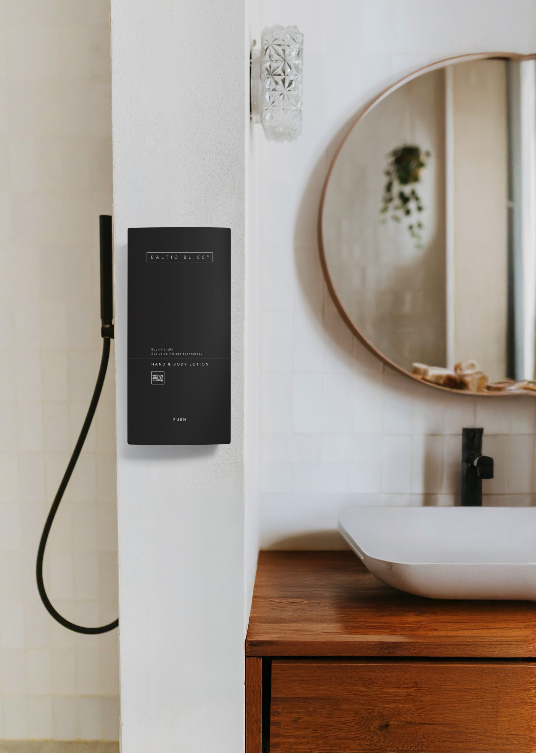 Een moderne badkamer met een zwarte wanddispenser voor hand- en bodylotion, naast een ronde spiegel en een witte wastafel op een houten aanrechtblad.