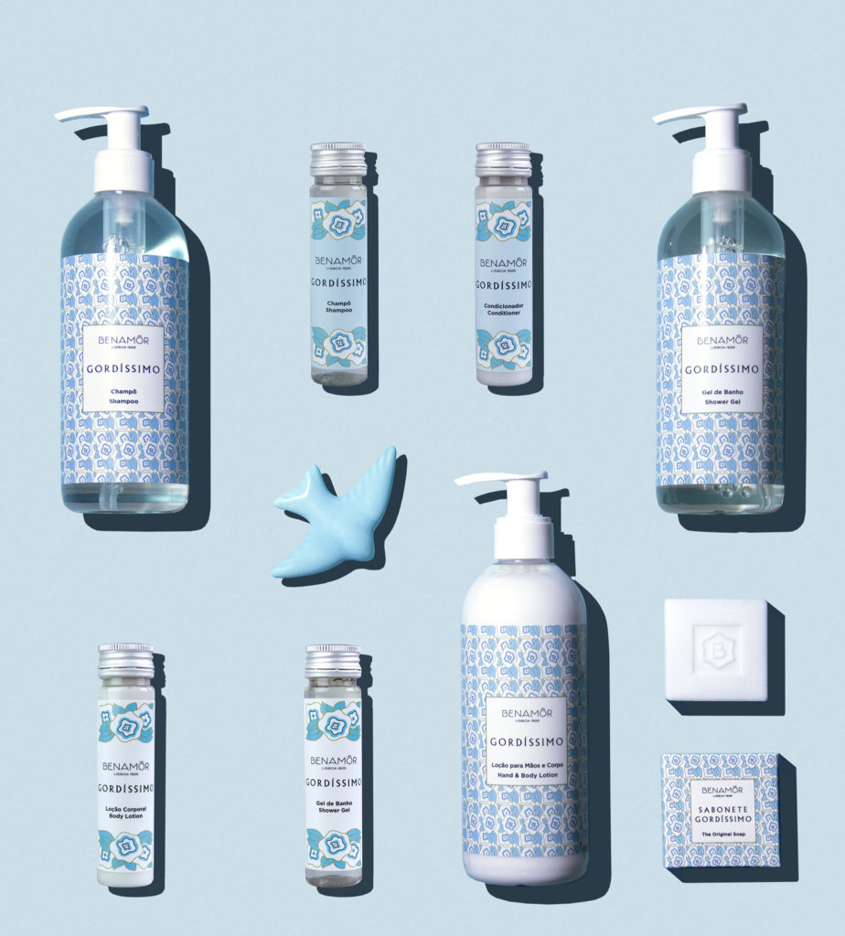 Una composizione piatta di vari prodotti per la cura della pelle del marchio "Denamorado Gordissimo", tra cui flaconi, barattoli e un sapone a forma di uccellino blu, disposti su uno sfondo azzurro. I prodotti sono caratterizzati da un'elegante confezione blu e bianca.