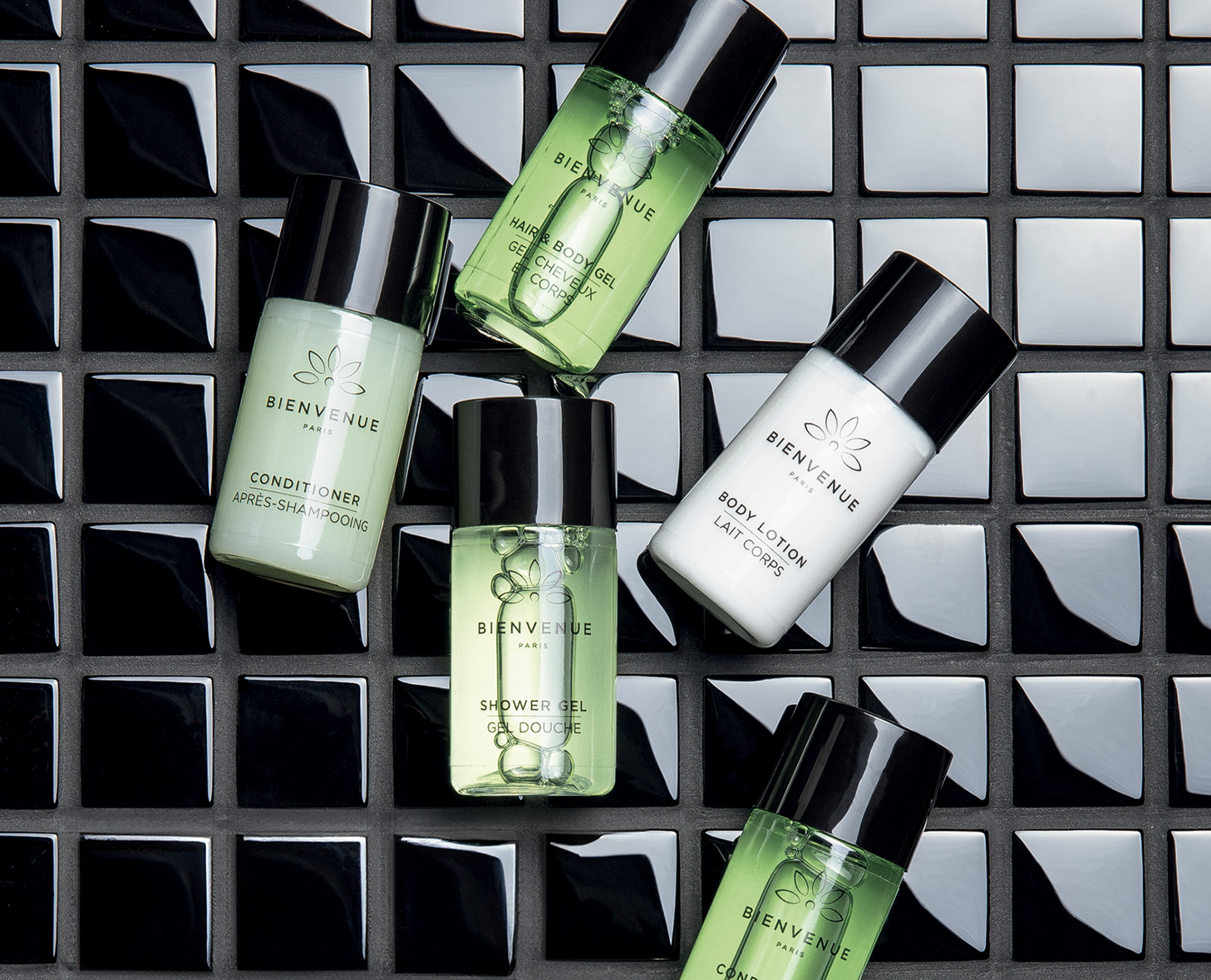 Eine Sammlung kleiner grüner und weißer Flaschen mit der AufschriftBIENVENUE“, darunter Conditioner, Haargel, Duschgel, Körperlotion und Shampoo, angeordnet auf einer schwarzen Fliesenoberfläche.