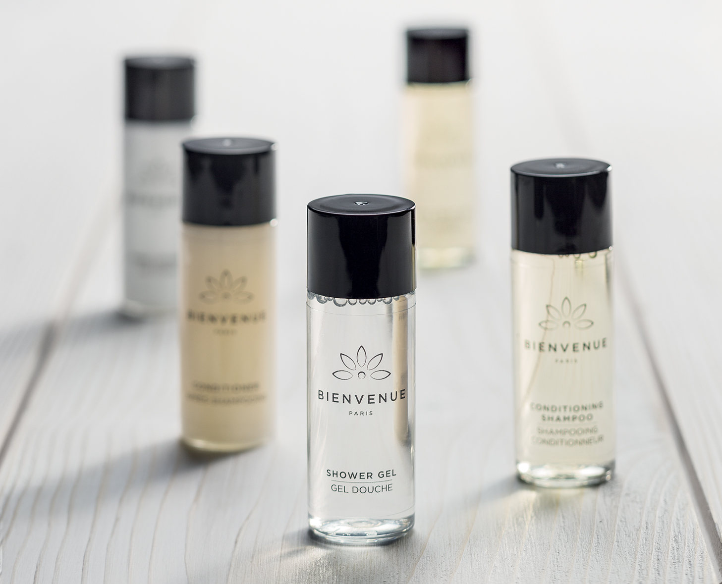 Eine Kollektion kleiner Flaschen mit Duschgel, Pflegeshampoo und anderen Körperpflegeprodukten, präsentiert auf einer hellen Holzfläche. Die Flaschen haben ein minimalistisches Design mit schwarzen Verschlüssen und dem auffällig platzierten MarkennamenBIENVENUE“.