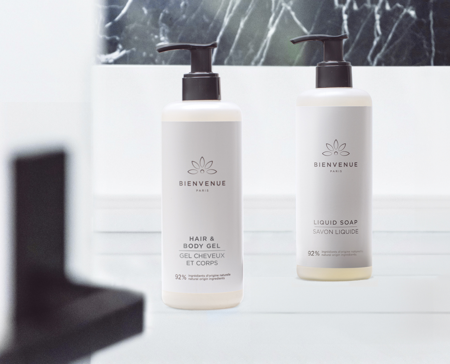 Zwei Flaschen mit Körperpflegeprodukten Bienvenue sind vor einem Marmorhintergrund ausgestellt: eine mit der Aufschrift „Hair &amp; Body Gel” und die andere mit „Liquid Soap”. Beide Flaschen haben ein minimalistisches Design mit einer hellen Farbgebung.