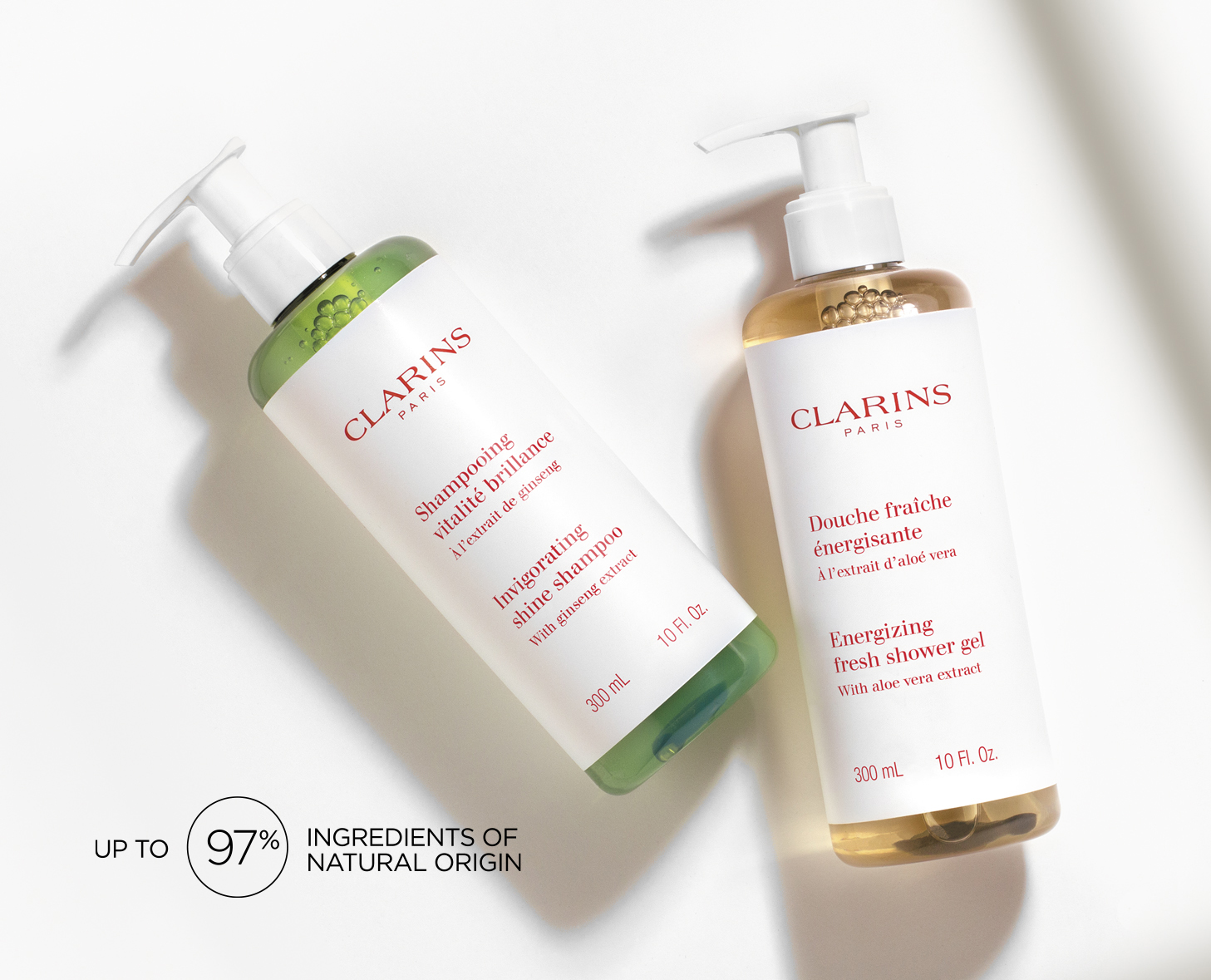 Zwei Haar- und Körperpflegeprodukte Clarins : eine grüne Flasche mit revitalisierendem Shampoo und eine orangefarbene Flasche mit energiespendendem Duschgel, beide mit Pumpspender, vor hellem Hintergrund.
