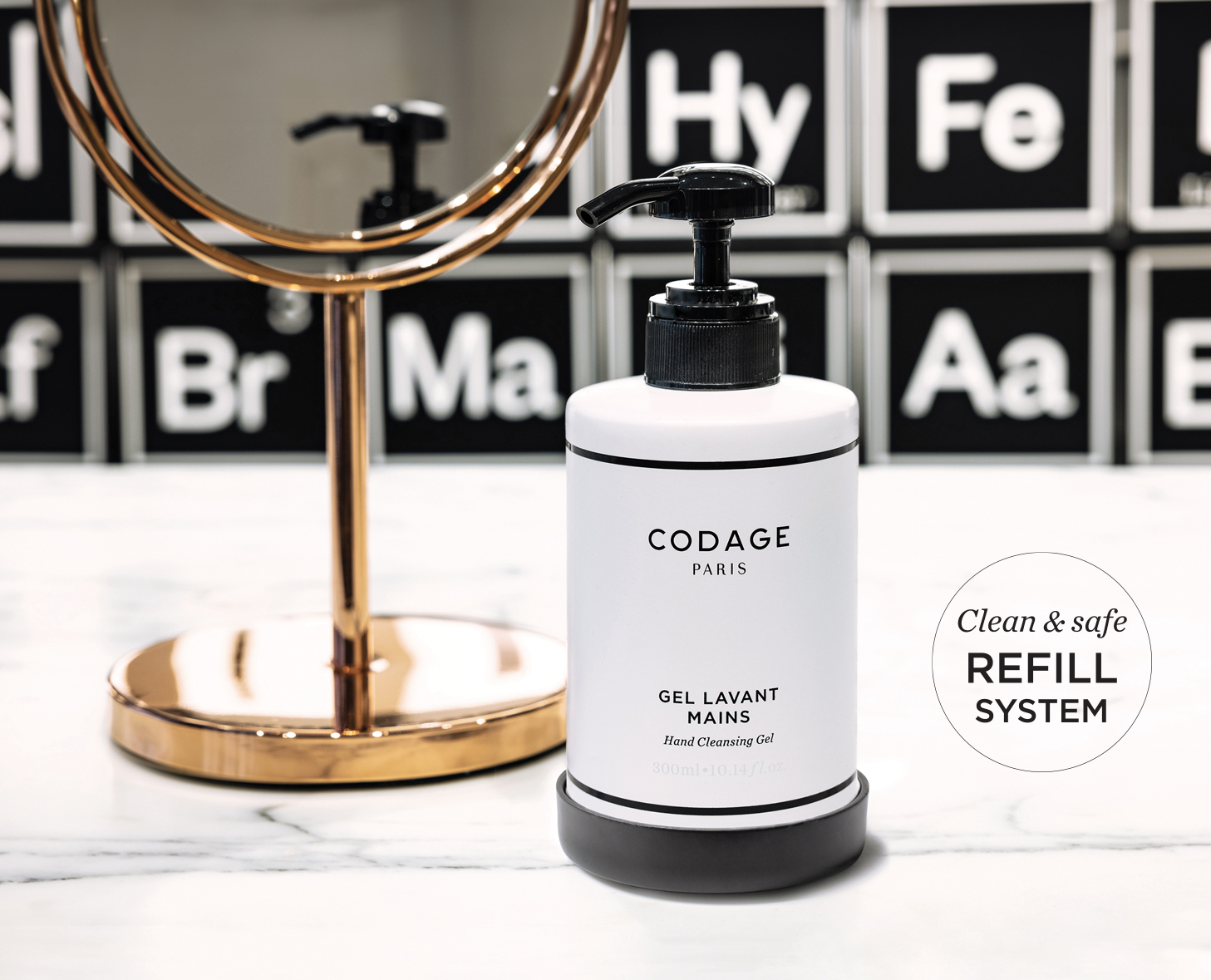 Eine Flasche CODAGE Handreinigungsgel steht auf einer Marmorplatte neben einem runden Spiegel, im Hintergrund sind Elemente des Periodensystems zu sehen. Das Etikett hebt ein sauberes und sicheres Nachfüllsystem hervor.