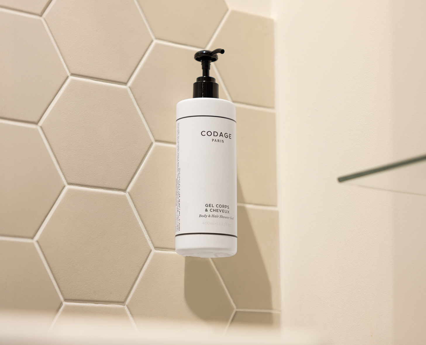 Eine weiße Flasche Codage gel mit der Aufschrift „Gel Corps &amp; Cheveux” ist an einer hellen, sechseckigen Fliesenwand in einem Badezimmer angebracht.