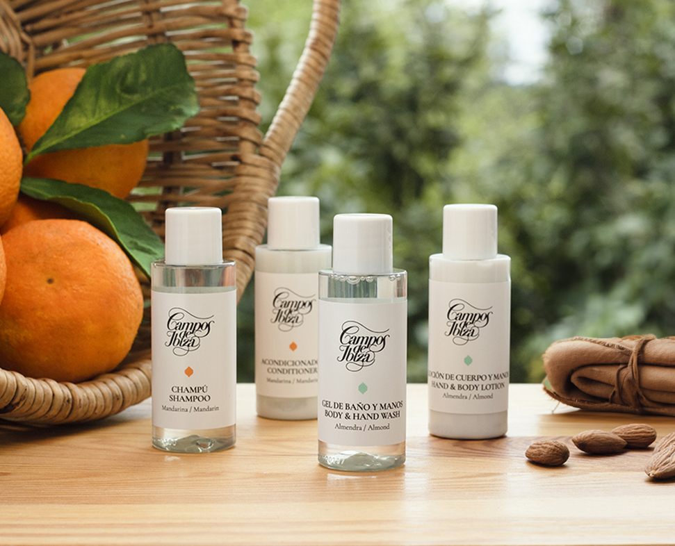 Quattro flaconi di prodotti per la cura personale, tra cui shampoo, balsamo, bagnoschiuma e lozione, sono esposti su una superficie di legno accanto a un cesto di arance e mandorle, sullo sfondo di un paesaggio naturale all'aperto.