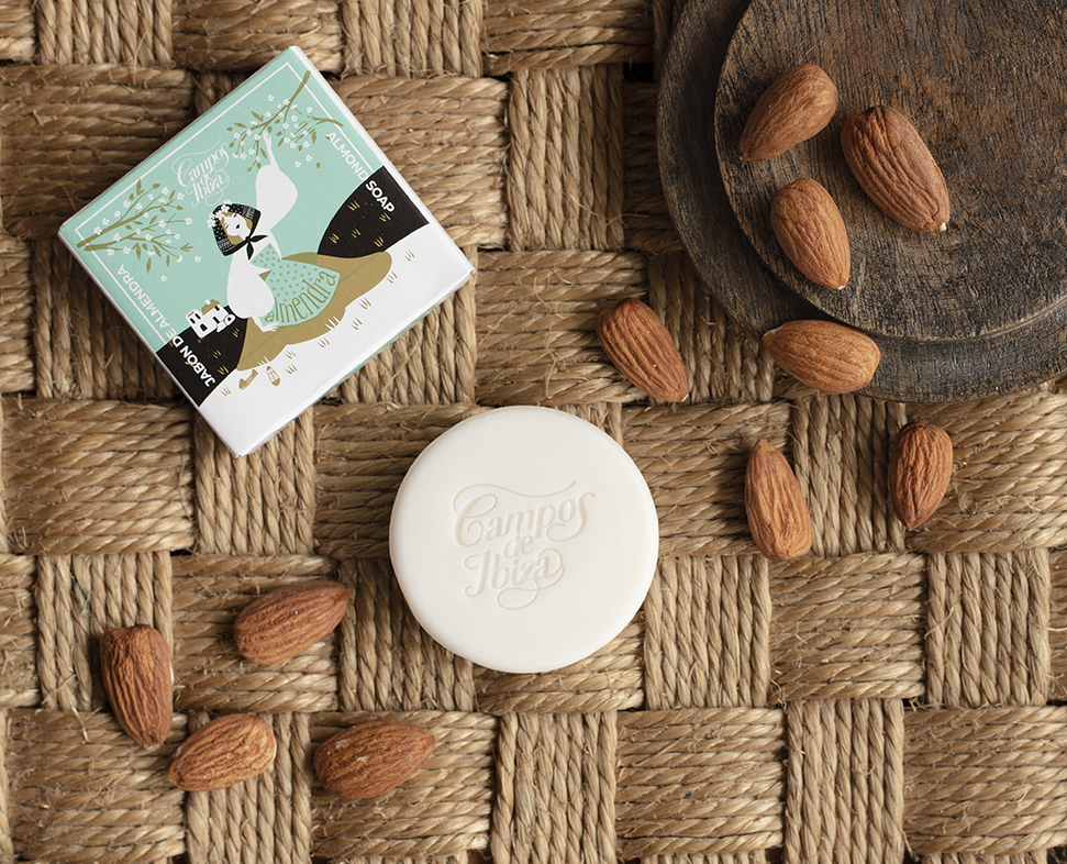 Una confezione decorativa di sapone profumato "Campos de Ibiza" è posizionata su una stuoia intrecciata, circondata da mandorle sparse.