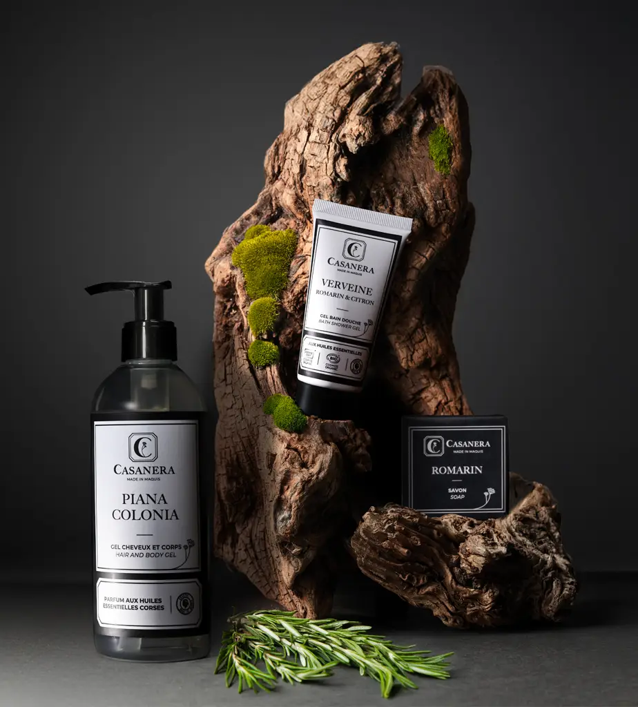 Auf einer dunklen, strukturierten Holzoberfläche sind drei Casanera ausgestellt: eine große Flasche Piana Colonia Duschgel, eine Tube Verveine Handcreme und ein Stück Romarin Seife, begleitet von einem Hauch von grünem Moos und Rosmarin.