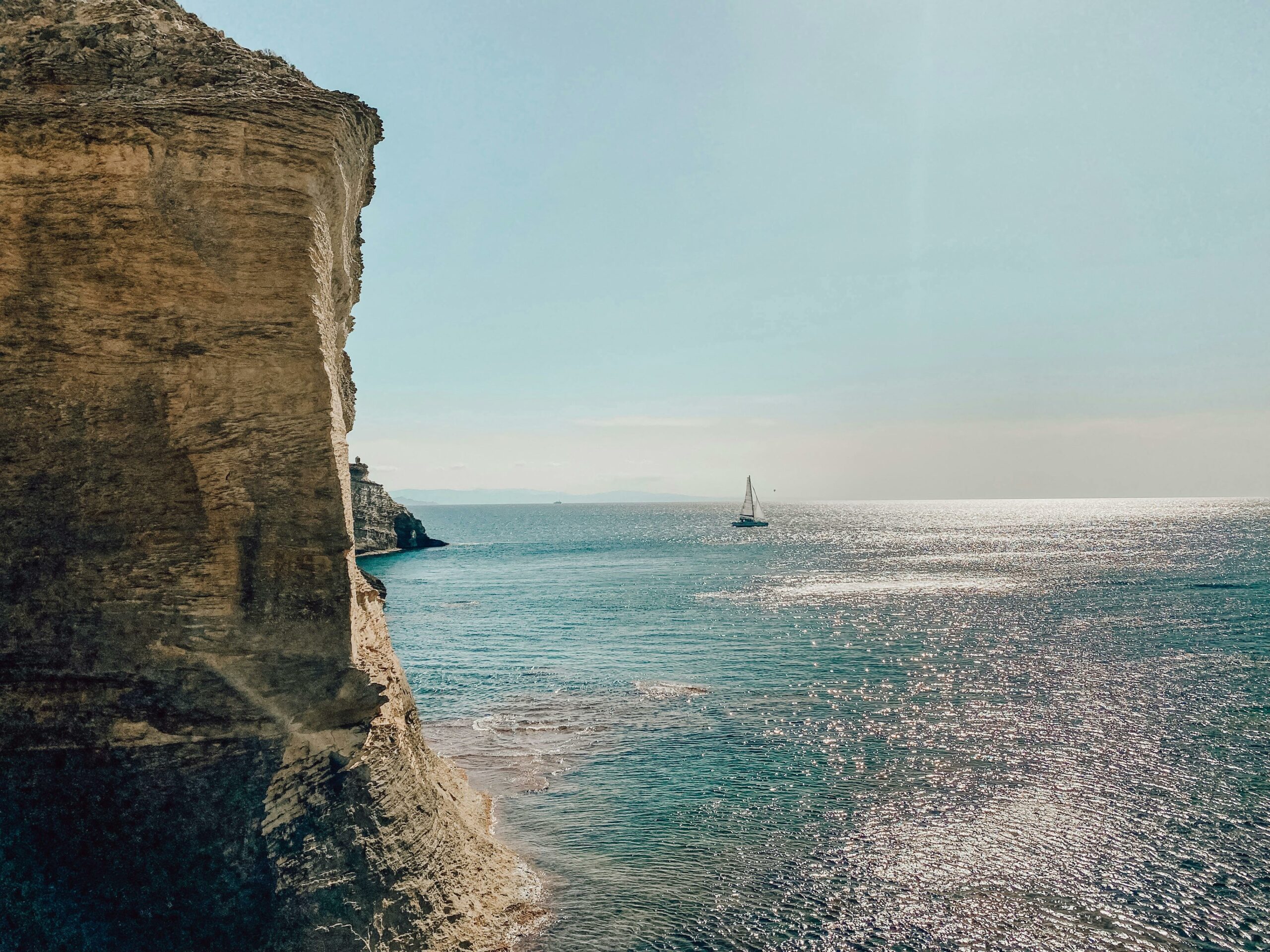 Una barca a vela scivola sulle acque blu scintillanti vicino a una scogliera frastagliata sotto un cielo luminoso. La luce del sole si riflette sull'oceano, creando un effetto scintillante.